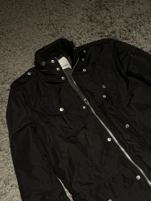 Svart field jacket från J.Lindeberg - Svart field jacket från J.Lindeberg med hög krage, silverfärgad dragkedja och tryckknappar. Fyra stora fickor framtill och en diskret logga på ärmen. Jackan har en clean och sportig look, perfekt för dig som gillar stilrena detaljer.