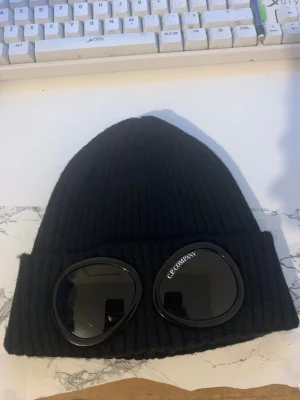 Svart ribbad goggla-mössa C.P. Company - Svart ribbstickad mössa från C.P. Company med ikoniska inbyggda goggles framtill. Klassisk uppvikt modell med tydlig logga på ena glaset. Perfekt accessoar för att sticka ut i vinterkylan och ge din outfit en streetwear-vibe.