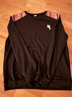 Svart sweatshirt med rutiga axlar Shein - Svart sweatshirt från Shein med rutiga detaljer i brunt och rosa på axlarna. Liten vit brodyr på bröstet. Klassisk rund halsringning och långa ärmar. Mjuk och skön, perfekt för en chill look.