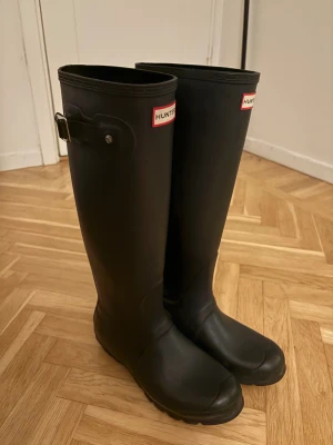 Svarta höga gummistövlar från Hunter - Klassiska svarta gummistövlar från Hunter med högt skaft och  spänne på sidan. Stövlarna har rund tå, platt sula och tydlig Hunter-logga framtill. Lite slitna på insidan men ger ett fint helhets intryck, se bilder 🩷