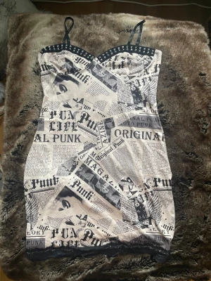 Kort punkig klänning med tidningsmönster - Säljer en kort klänning med svartvitt tidningsmönster och punkiga detaljer. Klänningen har smala axelband, nitar längs bysten och spetskant nertill. Perfekt för dig som gillar edgy och unik stil. Materialet känns mjukt och stretchigt, troligen polyester.