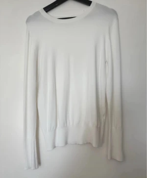 Vit långärmad stickad tröja med knappar - Vit stickad tröja med rund hals och långa ärmar. Ärmsluten har snygga guldfärgade knappar som ger en extra detalj. Tröjan har en mjuk och skön känsla, perfekt för lager-på-lager. Passar dig som gillar stilrena och enkla plagg med en twist. Använd Max 3 gånger, tyvärr uppkom snabbt noppror som det gör på sådan material, inte mycket och går nog att få bort syns på bild. En knapp har trillat av på ena ärmen!  