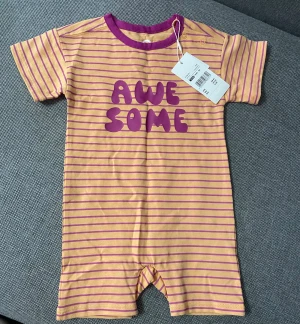 Randiga shortsdräkt med lila text - Säljer en kortärmad shortsdräkt från Lindex i ljusgul med lila ränder och lila texten 'AWESOME' framtill. Rund hals med lila kant och tryckknappar vid axeln. Perfekt för chill dagar.