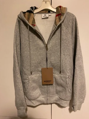 Grå hoodie från Burberry med rutig huva - Grå hoodie från Burberry med dragkedja och snörning. Insidan av huvan har klassiskt Burberry-rutigt mönster i beige, svart och rött. Tröjan har två fickor fram och ribbade muddar vid ärmslut och nederkant. Perfekt för chill dagar och snygg streetstyle.