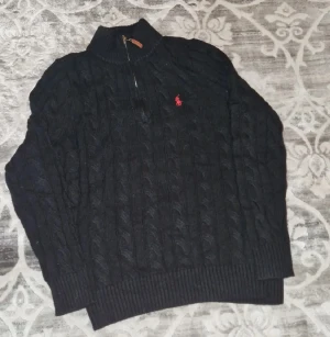 Svart stickad tröja Polo Ralph Lauren - Svart kabelstickad tröja från Polo Ralph Lauren med röd broderad logga på bröstet. Tröjan har half zip med läderdetalj på dragkedjan och ribbade muddar. Perfekt för dig som gillar klassisk stil med snygga detaljer.