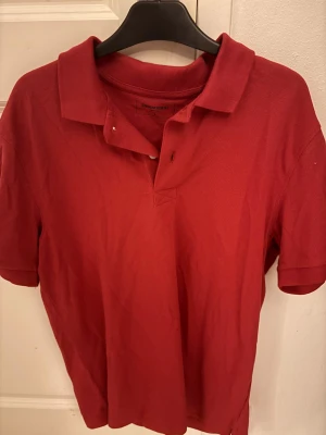 Röd pikétröja från Dressmann - Klassisk röd pikétröja från Dressmann med krage och korta ärmar. Tröjan har en enkel design med knappslå vid halsen och är tillverkad i ett bekvämt bomullsmaterial. Perfekt för en avslappnad och stilren look.