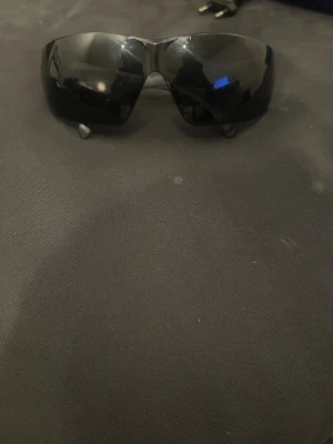 Svarta sportiga solglasögon från 3M - Säljer ett par svarta solglasögon från 3M med breda, svepande linser och snygga detaljer på skalmarna. Glasögonen har en sportig och futuristisk vibe, tillverkade i plast med mörka spegelglas. Perfekt för dig som gillar att sticka ut.