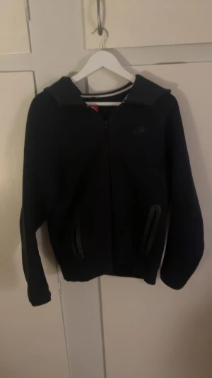 Svart hoodie från Nike med dragkedja - Svart hoodie från Nike med hel dragkedja framtill och två fickor med dragkedja. Klassisk huva och diskret Nike-logga på bröstet. Perfekt för chill eller träning, med långärmad passform och mjukt material.