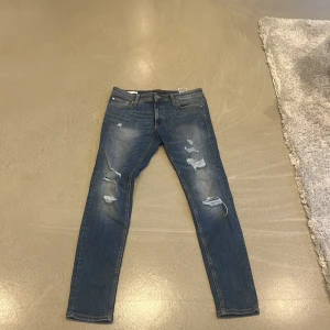 Blå skinny jeans Jack & Jones 32/32 - Säljer ett par blå skinny jeans från Jack & Jones i modellen LIAN. Jeansen har slitna detaljer och hål på benen för en cool, avslappnad look. Klassisk femficksmodell med dragkedja och knapp. Perfekta för dig som gillar en smal passform.