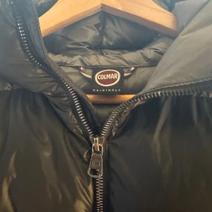 Svart pufferjacka från Colmar - Svart pufferjacka från Colmar Originals med huva och grov dragkedja framtill. Jackan har quiltad design och två sidofickor med dragkedja. Perfekt för kalla dagar och har en riktigt schysst, puffig look som är både trendig och bekväm. Den är i storlek 16/172cm men passar xs. Jackan är i nyskick!