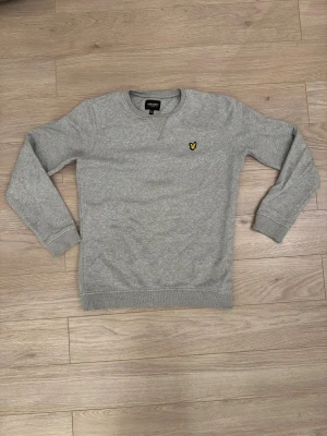 Grå sweatshirt från Lyle & Scott - Snygg grå sweatshirt från Lyle & Scott Junior med klassisk rund hals och långärmad design. Tröjan har det ikoniska gula fågelmärket broderat på bröstet och ribbade muddar vid ärmslut och nederkant. Perfekt för chill dagar och enkel att matcha.