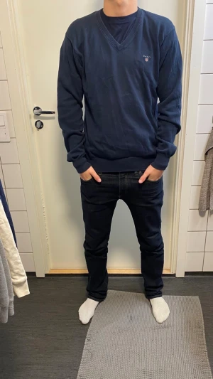 Mörkblå Gant tröja med mockadetaljer - Snygg mörkblå tröja från Gant med v-ringning och brunt mockalapp på armbågarna. Klassisk passform och diskret logga på bröstet. Perfekt till jeans och funkar året runt. Tillverkad i mjuk bomull för skön känsla.