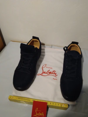 Christian Louboutin blåa sneakers - Snygga blåa sneakers från Christian Louboutin med klassisk röd sula och diskret broderad logga på hälen. Skorna har snörning, rund tå och är tillverkade i mocka med läderdetaljer. Perfekta för dig som vill ha en exklusiv och stilren look.
