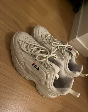 Vita chunky sneakers från Fila - Säljer ett par vita chunky sneakers från Fila med klassisk logga på sidan och plösen. Skorna har snörning, platt sula och är tillverkade i syntetmaterial. Perfekta för dig som gillar streetstyle och vill ha en sportig look.