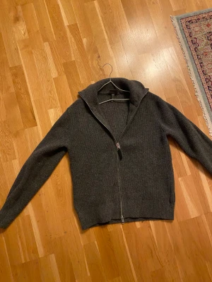 Mörkgrå stickad kofta med dragkedja - Mysig mörkgrå stickad kofta med hel dragkedja framtill och hög krage. Perfekt för kyliga dagar och enkel att matcha Flätat bälte i svart, rött och vittmed jeans eller chinos. Materialet är mjukt och ribbstickat, vilket ger en skön känsla och snygg struktur. Långärmad modell med normal passform.