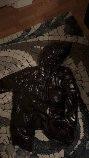 Svart Moncler pufferjacka med huva - Säljer en svart glansig pufferjacka från Moncler med huva och dragkedja framtill. Jackan har Moncler-logga på ärmen och flera praktiska fickor med dragkedja. Perfekt för kalla dagar när du vill ha både stil och värme.