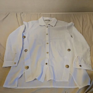 Vit skjorta med stora knappar  - Snygg vit skjorta med stora, silvriga knappar längs framsidan och på ärmsluten. Skjortan har klassisk krage, lång ärm och en loose passform. Helt oanvänd!