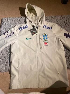 Vit Nike Brasil regnjacka L - Snygg vit regnjacka från Nike med Brasil-logga och sponsormärken. Jackan har huva, hel dragkedja och är tillverkad i vattentätt GORE-TEX-material. Perfekt för dig som vill ha sportig stil och hålla dig torr. Stora tryck på ärmar och bröst, meshfoder på insidan.