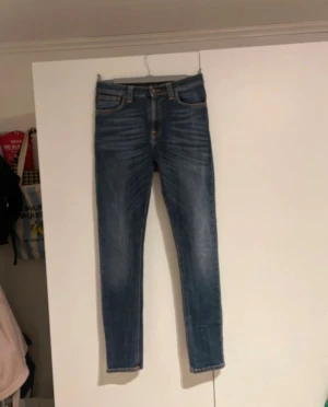Blå jeans från Nudie Jeans - Säljer ett par blå jeans från Nudie Jeans med klassiska orange sömmar och snygga slitningar. Jeansen har fem fickor, normal midja och är tillverkade i stretchig denim.