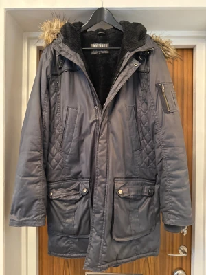 Grå parkas från Jofama med fuskpäls - Säljer en grå parkas från Jofama med huva och fuskpälsdetalj. Jackan har quiltade partier, flera fickor med knappar och dragkedja framtill. Insidan är fodrad med svart fleece för extra värme. Perfekt för kalla dagar och har en loose passform.