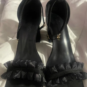 Svarta sandaler med volang och klack - Svarta sandaler med fyrkantig tå, klack och justerbar rem runt vristen. Framtill finns en snygg volangdetalj i organza som ger en extra touch. Skorna har guldfärgade spännen och är tillverkade i mockaliknande material. Perfekta för dig som gillar stilrena men ändå unika skor.