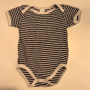 Randig kortärmad body i bomull - Kortärmad body i svart och vitt med smala ränder. Tillverkad i 100% bomull, med vita kantband runt hals, ärmar och ben. Praktisk knäppning med tryckknappar nertill. Perfekt för en chill och stilren look till de minsta.