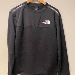 Svart tröja från The North Face  - Svart långärmad tröja från The North Face i storlek M. Tröjan har en sportig look med logga på bröstet och axlarna, samt en mix av slät och strukturerad polyester. Perfekt för lager-på-lager och chill vardagsstil.         Passar som s. 
