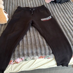 Svarta mjukisbyxor från Hoodrich M - Svarta mjukisbyxor från Hoodrich i storlek M. Byxorna har snörning i midjan och tryckt logga på ena benet. Tillverkade i mjukt material med resår vid bensluten för en skön och avslappnad passform.