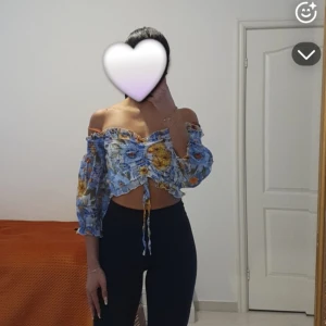 Blommig offshoulder croppad blus - Superfin blommig blus i blått, gult och vitt med offshoulder och trekvartsärmar. Toppen är croppad med snörning framtill och volangkant. Perfekt för sommarens alla tillfällen och ger en fräsch vibe.