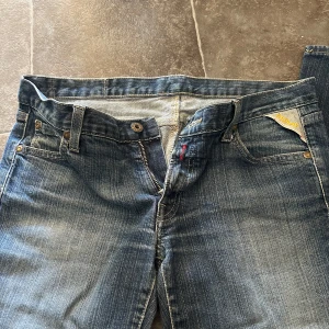 Blå Replay jeans med broderad patch - Snygga blå jeans från Replay med klassisk femficksdesign och dragkedja. Jeansen har en unik rosa patch med broderade blommor bak på linningen. Tillverkade i 98% bomull och 2% elastan för en skön passform.