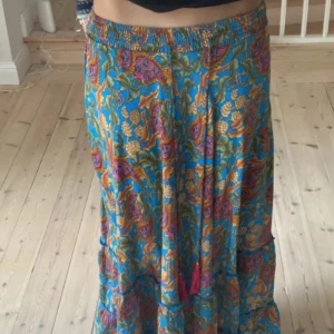 Blommig långkjol från Chelsea & Theodore - Färgglad långkjol med blommigt mönster från Chelsea & Theodore. Kjolen har elastisk midja och är i tunnt, flowy material. Basen är blå med inslag av grönt, gult, orange och rosa. Perfekt för dig som gillar boho-vibes och vill sticka ut med färg.