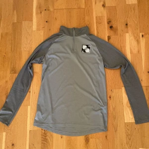 Grå half zip tröja från Under Armour - Sportig grå långärmad tröja från Under Armour med half zip och hög krage. Tillverkad i mjukt och lätt polyestermaterial, perfekt för träning. Tröjan har en lös passform och en stilren logga på bröstet.