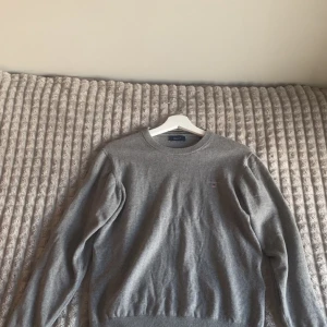 Grå stickad tröja från GANT - Klassisk grå stickad tröja från GANT med rund halsringning och broderad logga på bröstet. Tröjan har ribbade muddar vid ärmslut och nederkant, perfekt för lager-på-lager under höst och vinter.