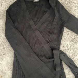 Svart omlottkofta från Urban Classics - Svart ribbad omlottkofta från Urban Classics i storlek XS. Koftan har långa ärmar, knytband i midjan och en snygg v-ringning. Perfekt för lager-på-lager och enkel att styla till olika looks.