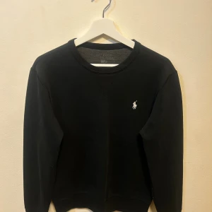 Svart sweatshirt från Polo Ralph Lauren - Klassisk svart sweatshirt från Polo Ralph Lauren med rund halsringning och broderad vit logga på bröstet. Tröjan har långa ärmar och ribbade muddar vid ärmslut och nederkant. Tröjan är i fint skick utan skador eller fläckar, färgen är fortfarande i fint skick trots att tröjan är några år gammal. Storlek M.