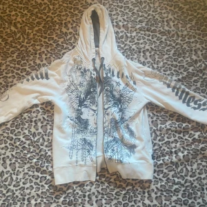 Vit hoodie med tryck  - Dragkedjan är sönder, går ej att knäppa!! Storlek XL med grymt tryck i svart på både fram- och baksidan. Tröjan har huva med snörning. Stora grafiska motiv och text längs ärmarna ger en edgy vibe. Perfekt för dig som gillar streetwear och vill sticka ut.