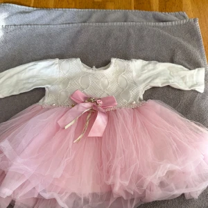Rosa och vit babyklänning med rosett - Supersöt babyklänning från Hulsen Baby i storlek 80-12M. Överdelen är vit med strukturmönster och pärldetaljer, medan kjolen är fluffig och rosa i flera lager tyll. Midjan pryds av en stor rosa rosett med guldband och små blommor. Klänningen har långärmade ärmar och rund hals.Rosa och vit babyklänning med rosett