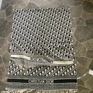 Svartvit halsduk från Christian Dior - Snygg halsduk från Christian Dior med svartvitt mönster och tydlig logga i båda ändar. Halsduken har fransar och ett grafiskt, rutig-inspirerat mönster med Dior-tryck över hela ytan. Perfekt för dig som gillar lyxiga accessoarer med statement.