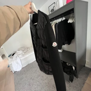 Svart cardigan från Moncler - Svart dunväst från Moncler med quiltad front och stickade svarta ärmar. Västen har rund hals, fyra fickor med knappstängning och Moncler-logga på ärmen. Materialet är en mix av dun och stickad textil, perfekt för kyliga dagar.