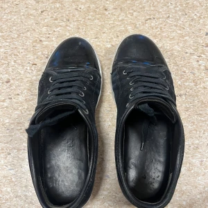 Svarta sneakers från Lanvin i mocka - Snygga svarta sneakers från Lanvin med ovandel i mocka och detaljer i blankt skinn på tån. Skorna har vita gummisulor och klassisk snörning med metallöljetter. Perfekta för dig som gillar stilrena och exklusiva sneakers med en lyxig känsla.