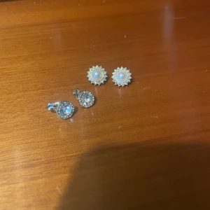 Två par glittriga örhängen - Säljer två par örhängen, ett par med vita pärlor omgivna av små glittrande stenar i solformad design och ett par med stora runda klara stenar i silverfärgad infattning. Perfekt för dig som gillar bling och klassisk stil. Dem är använda en gång var, annars fullt nya!