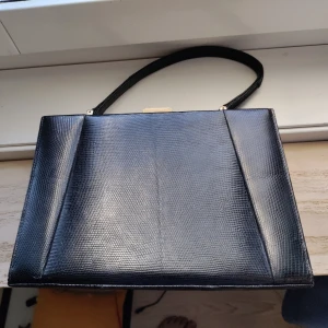 Väska - Det här är en snygg svart väska hårt material så den står upp av sig själv. Förutom det stora facket finns ett litet innerfack som öppnas med dragkedja. Det är inget särskilt märke. Storleken är 25x17x7 cm. 