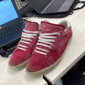 Rösa ysl skor - Säljer ett par röda sneakers i mocka med vita snören och beige sula. Skorna är tillverkade i Italien och har en klassisk, stilren look med rund tå och platt sula. Insidan är fodrad i ljust skinn för extra komfort. Perfekta för dig som gillar en unik touch. Skick 5/10 därav billigt pris. Dustbag följer med vid köp och priset är inte hugget i sten