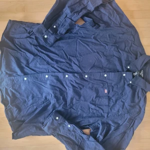 Mörkblå skjorta från Ralph Lauren - Snygg mörkblå skjorta från Ralph Lauren Jeans Co. i 100% bomull. Klassisk modell med knappar hela vägen, bröstficka med RL-detalj och lång ärm. Perfekt för dig som gillar stilrena och tidlösa plagg.