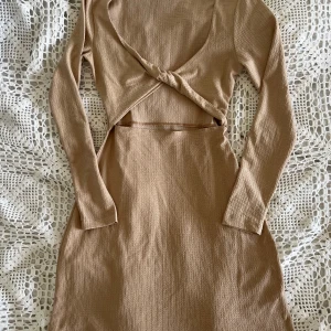 Långärmad klänning med twist-detalj  - Säljer en beige långärmad klänning från NLY med twistad detalj framtill och snygg cutout under bysten. Klänningen är ribbad och har en kroppsnära passform som framhäver siluetten. Materialet är mjuk polyester med lite stretch för extra komfort.
