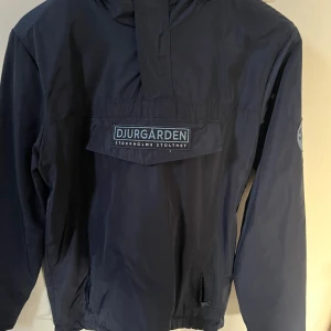 Djurgården vindjacka med huva - Mörkblå vindjacka från Djurgårdens IF med broderad DIF-logga på ärmen och stor ficka framtill med Djurgården-tryck. Jackan har huva, dragkedjor på fickorna och är tillverkad i lätt polyester. Perfekt för dig som vill visa vilket lag du håller på.