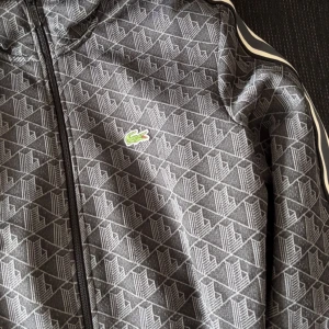 Grå mönstrad ziptröja från Lacoste - Snygg grå ziptröja från Lacoste med geometriskt mönster och klassisk krokodillogga på bröstet. Tröjan har svarta och vita ränder på axlarna och dragkedja framtill. Regular fit och perfekt för dig som gillar sportig och stilren look. Den passar mig inte längre men är ungefär oanvänd.