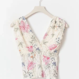 Blommig topp  - Romantisk linnetopp i vit spets med blommigt mönster i rosa, blått och grönt. Toppen har djup v-ringning och är ärmlös, perfekt för varma dagar. Den croppade modellen ger en trendig och feminin look.