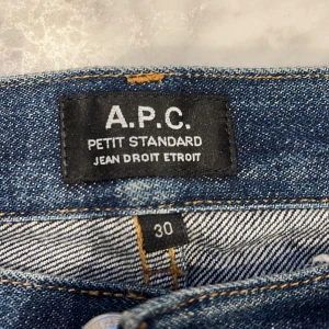 A.P.C. Petit Standard jeans blå 30 - Säljer ett par klassiska A.P.C. Petit Standard jeans i mörkblå denim. Modellen har raka ben, fem fickor och snygga slitningar vid knän och bak. Jeansen är tillverkade i bomull och har en stilren, tidlös look med orange kontrastsömmar.