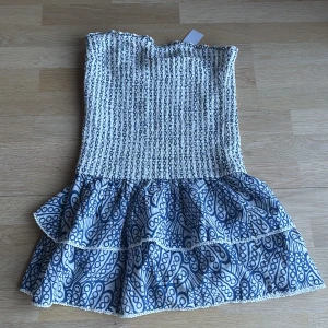 Vit och blå smockad kortklänning - Supersöt axelbandslös kortklänning med smockad överdel i vitt och blått. Kjolen har två volanger och ett blått abstrakt mönster. Klänningen är lätt och luftig, perfekt för varma dagar. Resår upptill för extra komfort och snygg passform.